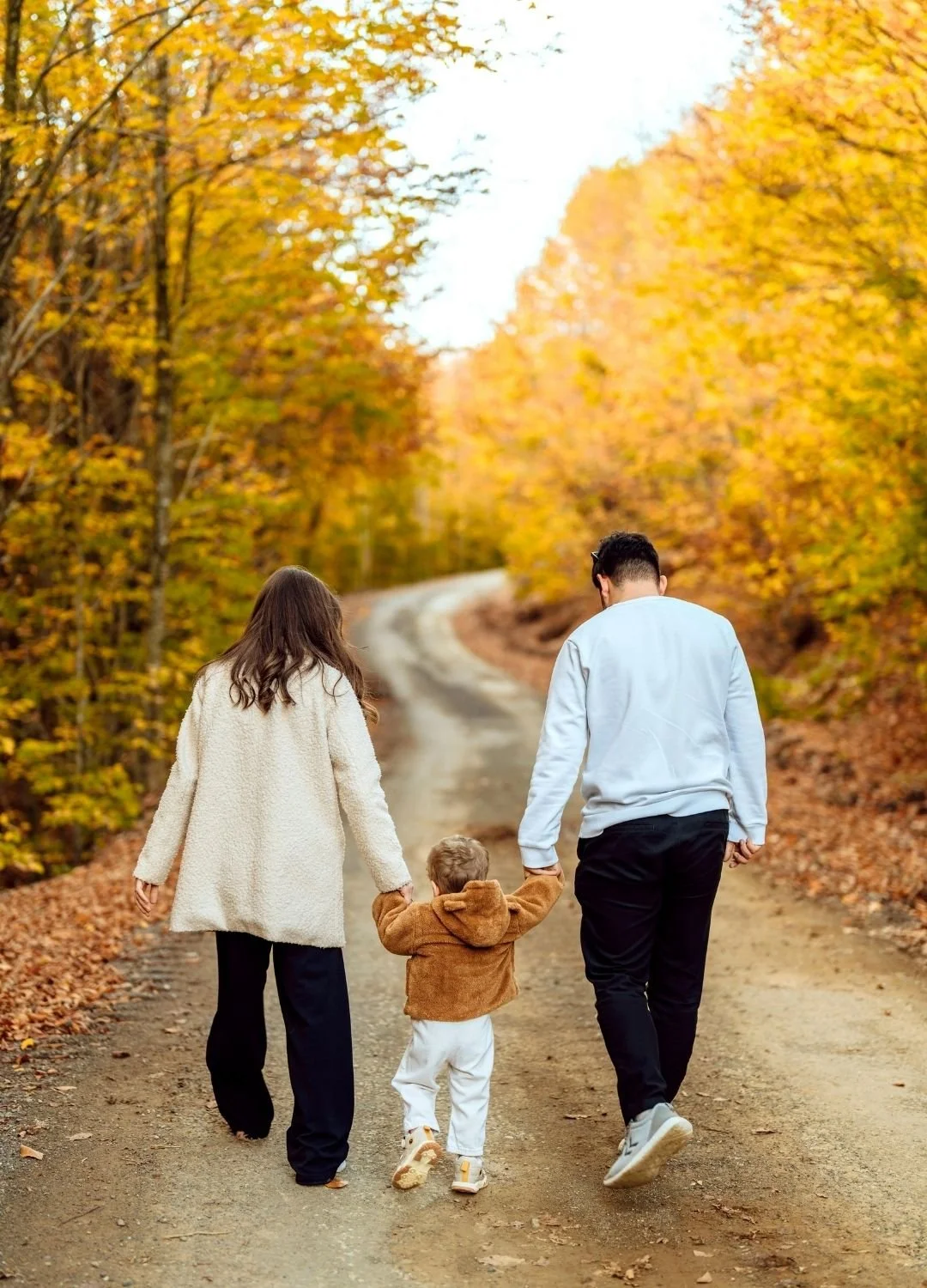 Two adults and a child walk hand in hand down a dirt road surrounded by trees with autumn foliage.