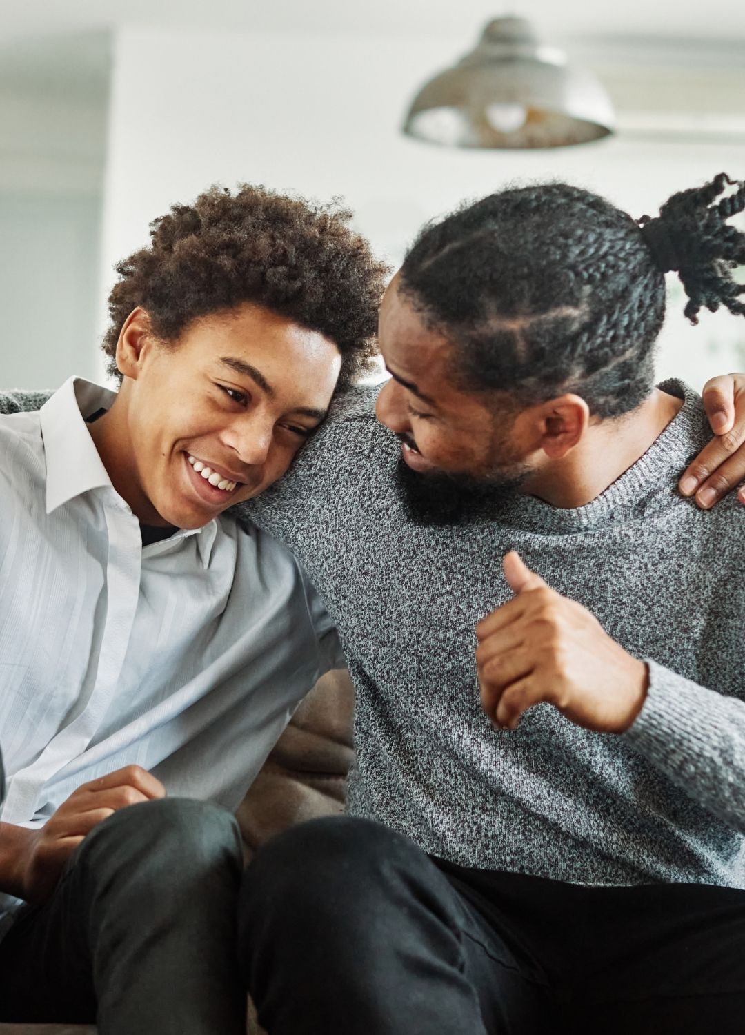 Two people sit close together on a couch, smiling and appearing to enjoy a friendly conversation. One has an arm around the other in a gesture of affection.