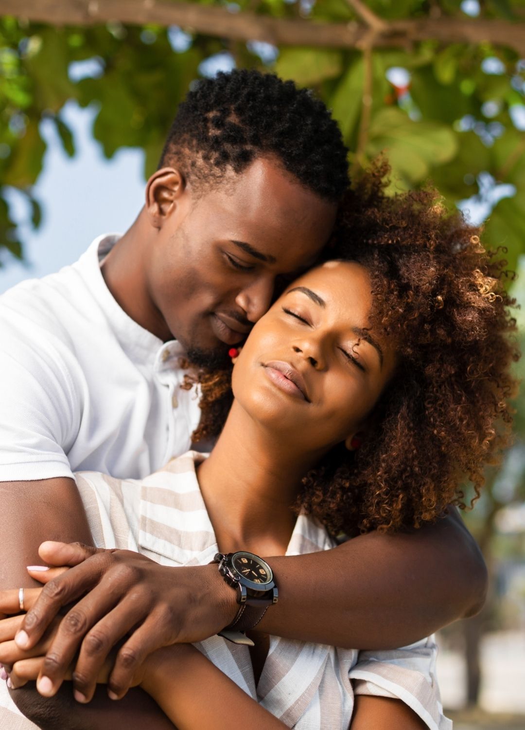 A man embraces a woman from behind outdoors; both appear relaxed, with the woman’s eyes closed and a calm expression.