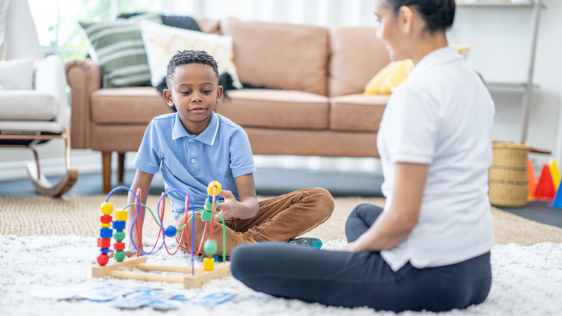 A child and an adult sit on a rug in a living room, interacting with a bead maze toy.