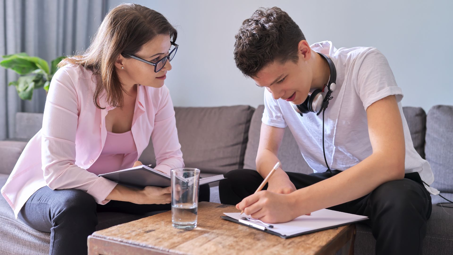 A woman watches as a teenage boy with headphones writes on a clipboard at a table.
