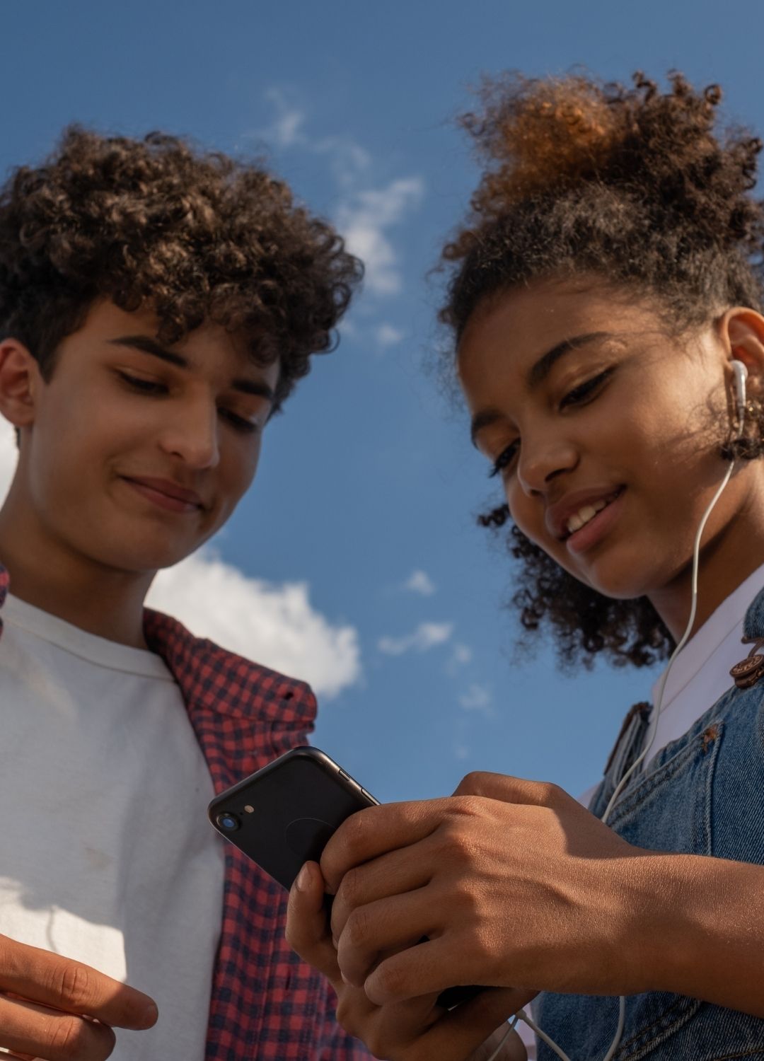 Two teenagers stand outdoors under a blue sky, looking at a smartphone together. One wears headphones.